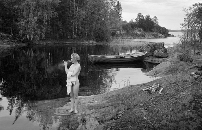 Mustavalkoinen valokuva, jossa nainen seisoo alastoman pyyhkeen kanssa rannalla tupakoimassa.